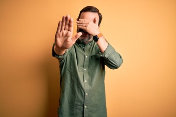 Middle age hoary man wearing casual green shirt and glasses over isolated yellow background covering eyes with hands and doing stop gesture with sad and fear expression. Embarrassed and negative