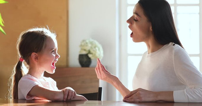 Caring skilled mother tutor teaching little preschool kid girl foreign language sounds pronunciation, sitting at table. Small child practicing articulation with therapist at private lesson at home.