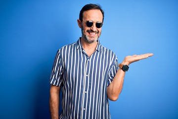 Naklejka premium Middle age hoary man wearing striped shirt and sunglasses over isolated blue background smiling cheerful presenting and pointing with palm of hand looking at the camera.