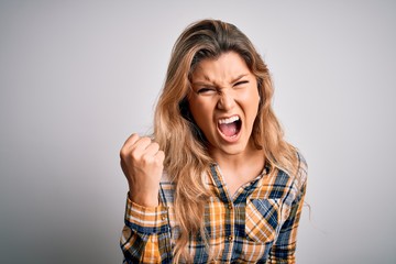 Young beautiful blonde woman wearing casual shirt standing over isolated white background angry and mad raising fist frustrated and furious while shouting with anger. Rage and aggressive concept.