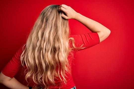 Young beautiful blonde woman wearing casual t-shirt standing over isolated red background Backwards thinking about doubt with hand on head