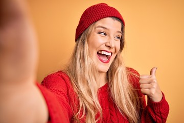Young beautiful blonde woman making selfie by camera over isolated yellow background pointing and showing with thumb up to the side with happy face smiling