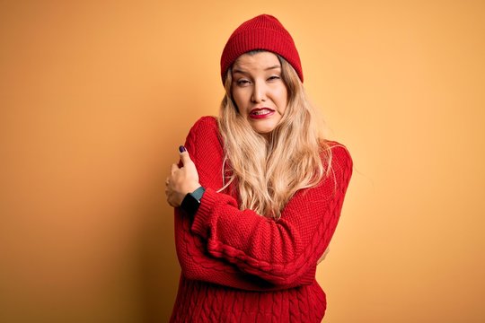 Young beautiful blonde woman wearing casual sweater and wool cap over white background shaking and freezing for winter cold with sad and shock expression on face