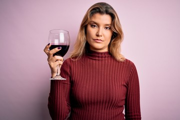 Young beautiful blonde woman drinking glasse of red wine over isolated pink background with a confident expression on smart face thinking serious