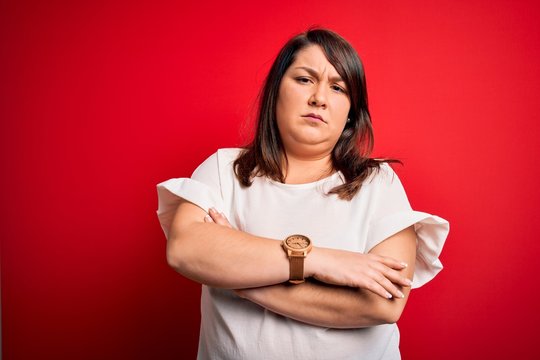 Beautiful brunette plus size woman wearing casual t-shirt over isolated red background skeptic and nervous, disapproving expression on face with crossed arms. Negative person.
