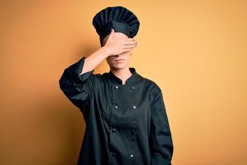 Young beautiful chef woman wearing cooker uniform and hat standing over yellow background covering eyes with hand, looking serious and sad. Sightless, hiding and rejection concept