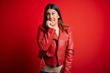 Young beautiful woman wearing red leather jacket over isolated background looking stressed and nervous with hands on mouth biting nails. Anxiety problem.