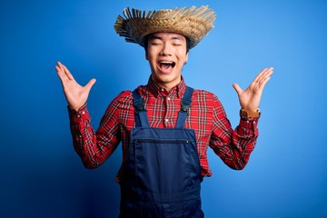 Young handsome chinese farmer man wearing apron and straw hat over blue background celebrating mad...