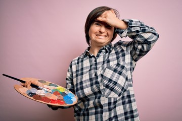 Young down syndrome artist woman holding painter palette and paintbrush over pink background...