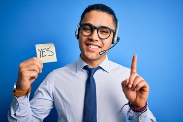 Young brazilian call center agent man holding reminder paper with yes message surprised with an...