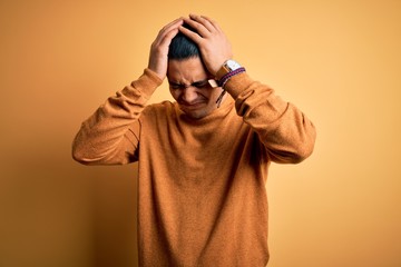Young brazilian man wearing casual sweater standing over isolated yellow background suffering from headache desperate and stressed because pain and migraine. Hands on head.