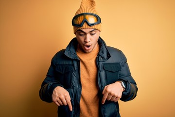 Young brazilian skier man wearing snow sportswear and ski goggles over yellow background Pointing down with fingers showing advertisement, surprised face and open mouth