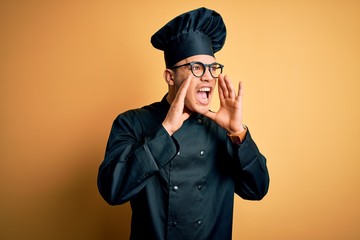 Young brazilian chef man wearing cooker uniform and hat over isolated yellow background Shouting angry out loud with hands over mouth