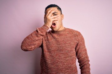 Young brazilian man wearing casual sweater standing over isolated pink background peeking in shock covering face and eyes with hand, looking through fingers with embarrassed expression.