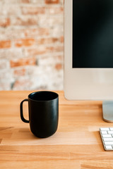 Image of a minimal desk setup with a small white keyboard, a computer monitor and a black coffee or tea mug on a wooden desk top