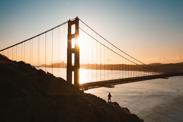A man standing alone in front of the famous Golden Gate Bridge at sunrise. Beautiful golden light behind the bridge. 