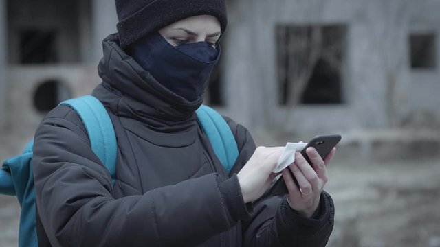 Young woman in a protective mask cleans her smartphone with sanitary napkin against the background of an abandoned building in an industrial post-apocalyptic landscape. Pandemic protection