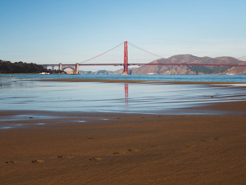 The famous Golden Gate Bridge shot at midday. The image of the bridge is reflected in the water on the sand. 
