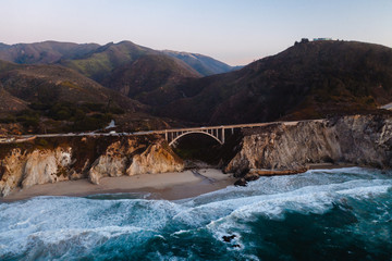 Big Creek Bridge in Big Sur, California. The photo is taken aerially from a drone as the sunsets in the distance, creating beautiful light on the bridge.