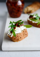 Italian bruschetta (sandwich, crostini, toast) with confit tomatoes, feta, olive oil and arugula leaves on white marble background. Traditional italian appetizer or snack, antipasto. Selective focus