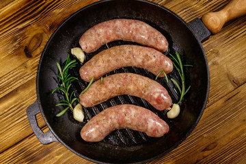 Raw sausages ready for preparation with rosemary, garlic and spices in cast iron grill frying pan on wooden table. Top view