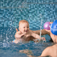 Fototapeta na wymiar Adorable cheerful child is splashing in blue water smiling and trying to reach for his ball in his mom's hands, cropped hands helping him to float