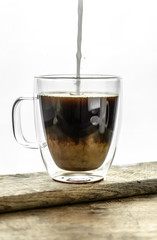 Pouring milk into the black coffee in transparent cup on wooden table