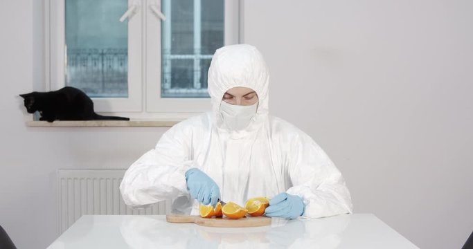 Portrait of young male person stitting behind white dining desk in kitchen in protective suit cutting an orange to eat later with a black cat in background during pandemic coronavirus quarantine time