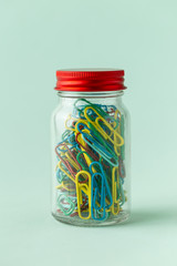 A small glass jar with a red lid full of colorful paperclips on a green background