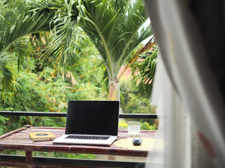 laptop of a remote digital nomad on a wooden bamboo table with notebook, mobile phone and glass in...
