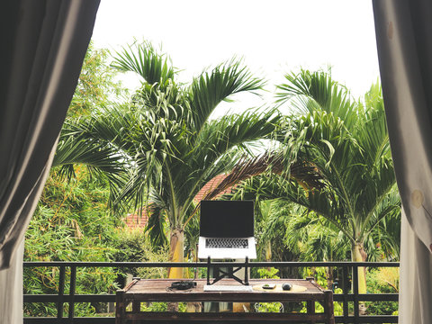laptop of a remote digital nomad on a wooden bamboo table with notebook, mobile phone, glass and chair in nature on a balcony with a green tropical background with palm trees