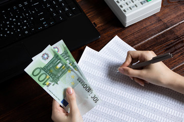 Obligation to pay wages and debts in the company.A cashier holds money Euro over an office work space with documents, a cash register,a phone,and a computer.Work in the office with finances in Europe