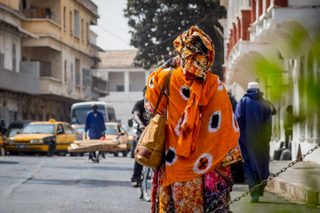 Back view of a typical african woman dressed in colorful  orange clothing on the streets of Sant Louis, Senegal during midday. - obrazy, fototapety, plakaty