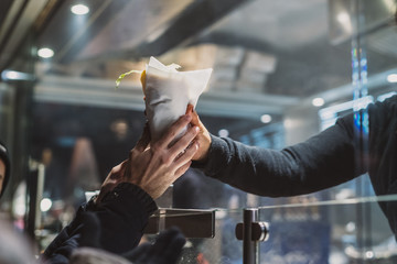 Person receiving a fresh kebab or gyros in a fast food stall or vendor outside in a city. Visible hands exchanging fast food over the counter in festive evening. - obrazy, fototapety, plakaty