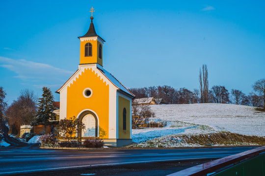 A small yellow church or a capel in Austria on a sunny winter time. Typical chapel in Austria or central part of europe lit by evening autumn sun. Some snow around on the hills, road leading past the 