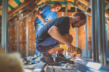 A person cutting holders, aluminium profiles or aluminium structural frames that hold drywall boards with an angle grinder in a house that is being rebuilt. Work on the roof using an angle grinder - obrazy, fototapety, plakaty
