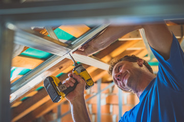 A young caucasian craftsman screwing tin holders for a drywall into a wood portion of a roof using...