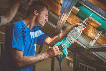 A person is cutting a board of wood next to a window with a reciprocating saw. Sawdust is flying...