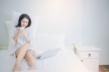 Happiness Asian woman with a cup of coffee start working day on a laptop sitting on the bed in the morning. Working from home concept