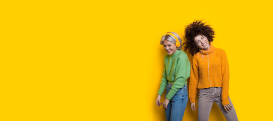 Curly haired friends listening to music using headphones on a yellow freespace while dancing happily