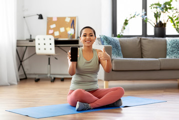 Fototapeta na wymiar sport, fitness and technology concept - happy smiling young african american woman sitting on exercise mat showing smartphone and tumbs up at home