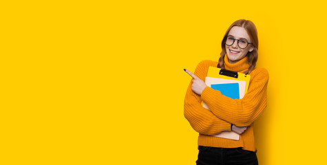 Ginger student with freckles and eyeglasses pointing to the yellow free space near her with finger embracing some books