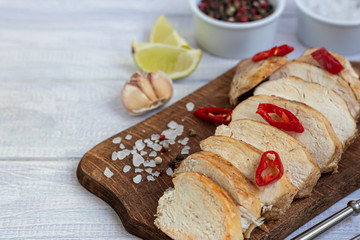 Cooked chicken breast in the oven, sliced on a wooden board. Served with chili pepper. Nearby are garlic, coarse salt, lime, peppercorns. Dietary nutrition.
