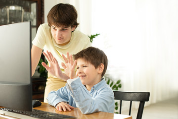 Smiling boy and teenager chatting online and waving at the computer screen. Quarantine and self-isolation due to coronavirus.