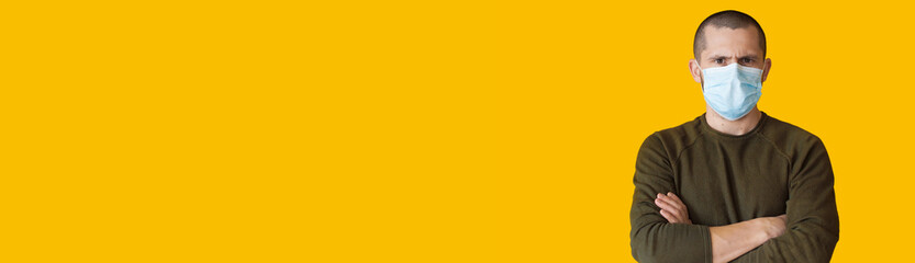 Sad caucasian man posing with crossed hands wearing medical mask on a yellow studio wall with free space