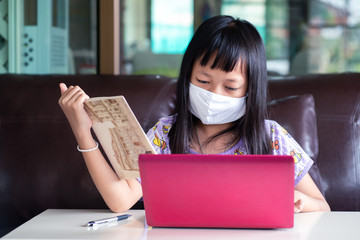 Asian child girl studying homework and wearing face mask during her online lesson at home for protect 2019-nCoV or Covid 19 virus , online education concept.