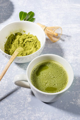 Matcha green tea in white cup, matcha green tea powder, special wooden spoon, bamboo whisk, fresh leaves on grey textured backdrop with shadows. Natural organic product with antioxidants for healthy.