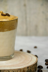 Vertical photo of a close up of whipped instant coffee with two coffee beans on a glass of milk on a wooden coaster with blurred background. Dalgona coffee concept