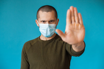 Serious caucasian man with short hair posing on a blue wall is gesturing the stop sign wearing a medical mask