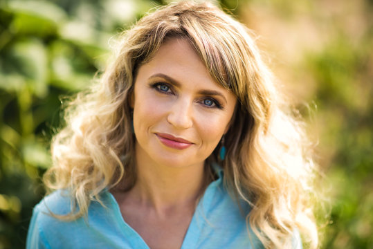 Close-up Portrait Of Beautiful Smiling Blonde 40 Years Old Woman In Blue Shirt Looking In Camera Outdoors In Back Light. Healthy Lifestyle And Shining Skin In Age Of 40s.